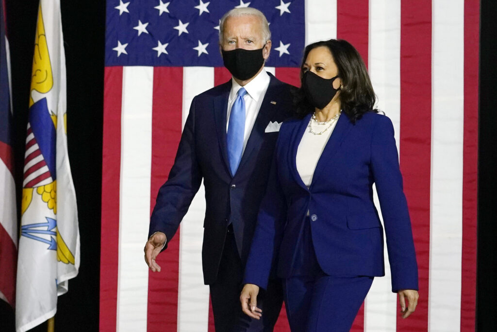 A man and woman wearing masks standing in front of an american flag.